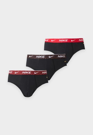 Млад мъж стои бос, облечен в черни боксерки Nike с оранжева лента на талията, позирайки пред бял фон.