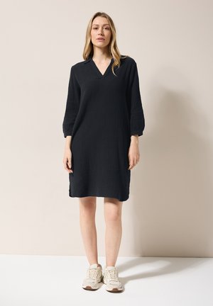 Robe en coton noire avec un col en V, des manches trois-quarts et une finition texturée. Associée à des baskets beiges.