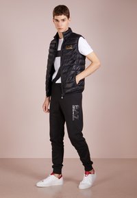 Must must vest, millel lukuga, valge ja musta triibuga T-särgi peal. Kombineeritud mustade joggerite ja punaste detailidega valgete tossudega.