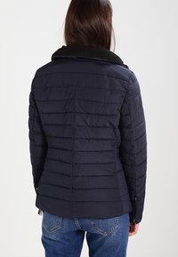 Kobieta ubrana w granatową pikowaną kurtkę z czarnym kołnierzem i niebieskie dżinsy, pokazana z tyłu na jednolitym tle.