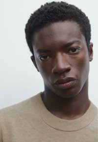 Close-up of a person with short, curly black hair wearing a light beige, knit sweater with a round neckline and ribbed edges.
