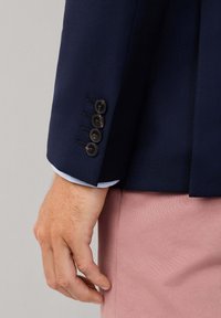 Manica di un blazer blu navy con quattro bottoni neri rotondi, tessuto testurizzato e un polsino di camicia azzurro chiaro che spunta sotto. Pantaloni rosa visibili.