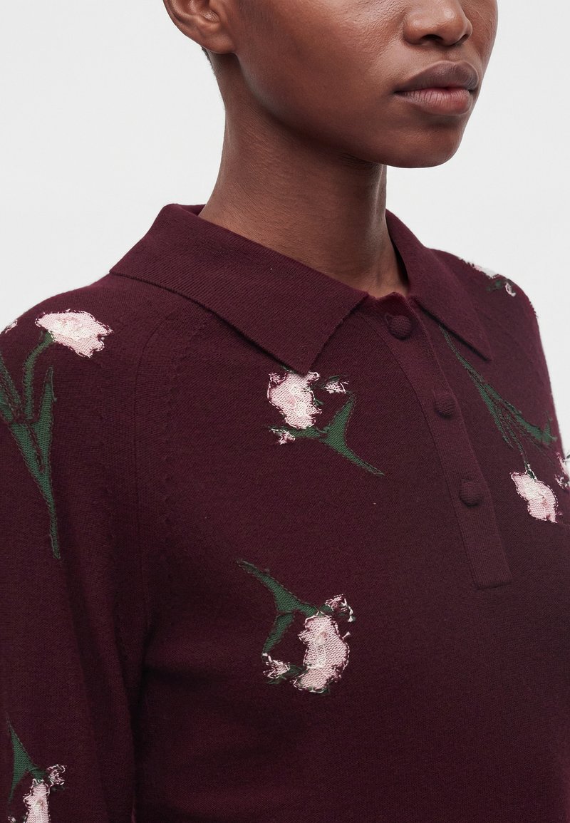 Person mit bordeauxfarbenem Pullover mit Kragen, hellrosa Blumenmuster und grünen Blättern, von den Schultern aufwärts vor einem schlichten Hintergrund.