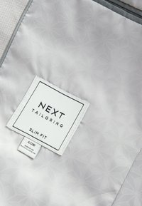 Étiquette de veste de costume blanche indiquant « NEXT TAILORING SLIM FIT » avec la taille 40R et « Made in Bangladesh » sur une doublure intérieure claire à motifs.