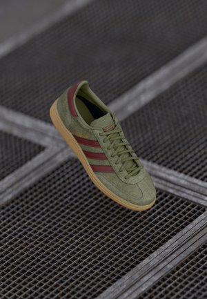 Grüner Wildleder-Adidas-Sneaker mit roten Streifen und Gummisohle, der auf einer dunklen Metallgitterfläche ruht.