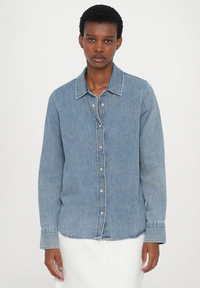 Denim shirt in light blue with long sleeves, button closure, pointed collar, and white stitching accents. Features a relaxed fit and textured fabric.