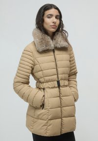 Donna che indossa una giacca trapuntata beige con collo in pelliccia, con le mani nelle tasche, in piedi davanti a uno sfondo grigio chiaro uniforme.