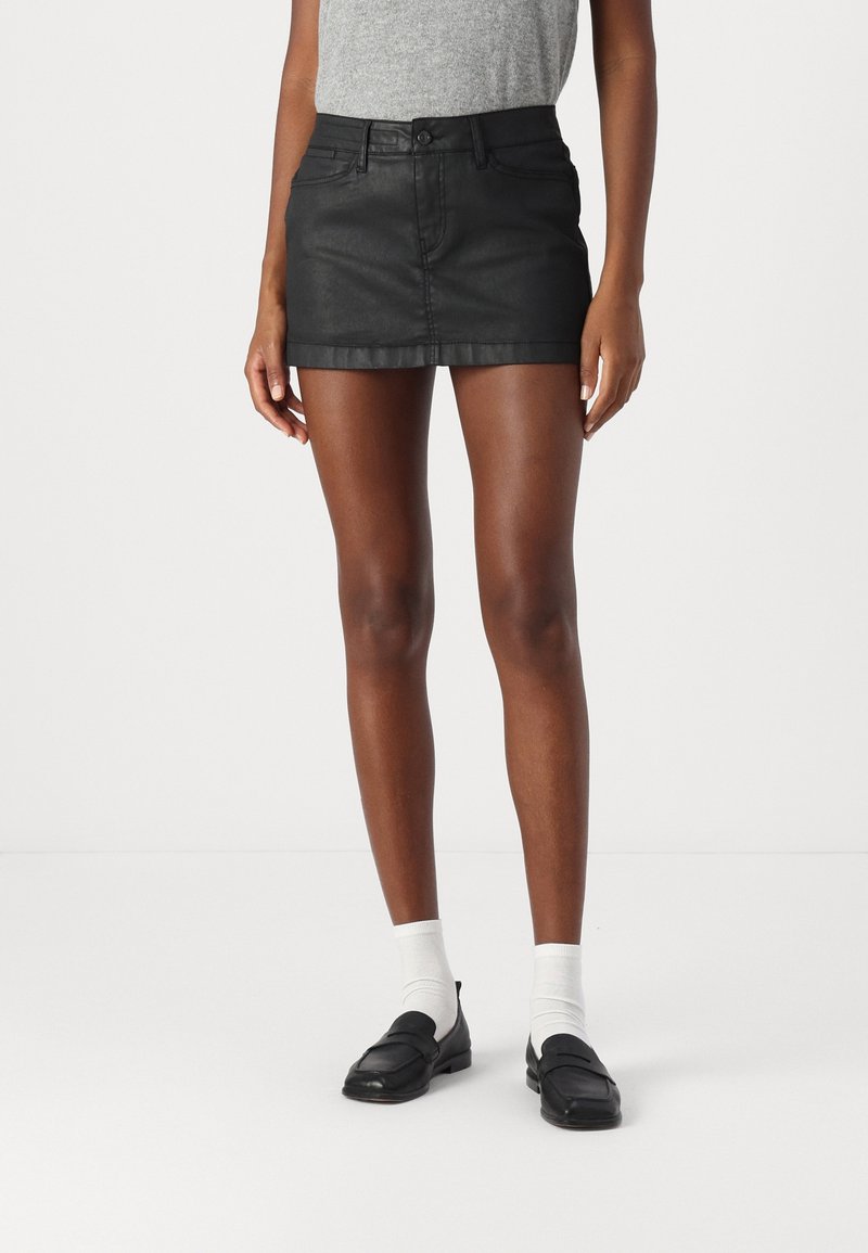 Černá džínová mini sukně s přiléhavým střihem, zapínáním na knoflík vpředu a pěti kapsami. Hladká textura, rovný lem, nošena s bílými ponožkami a mokasíny.