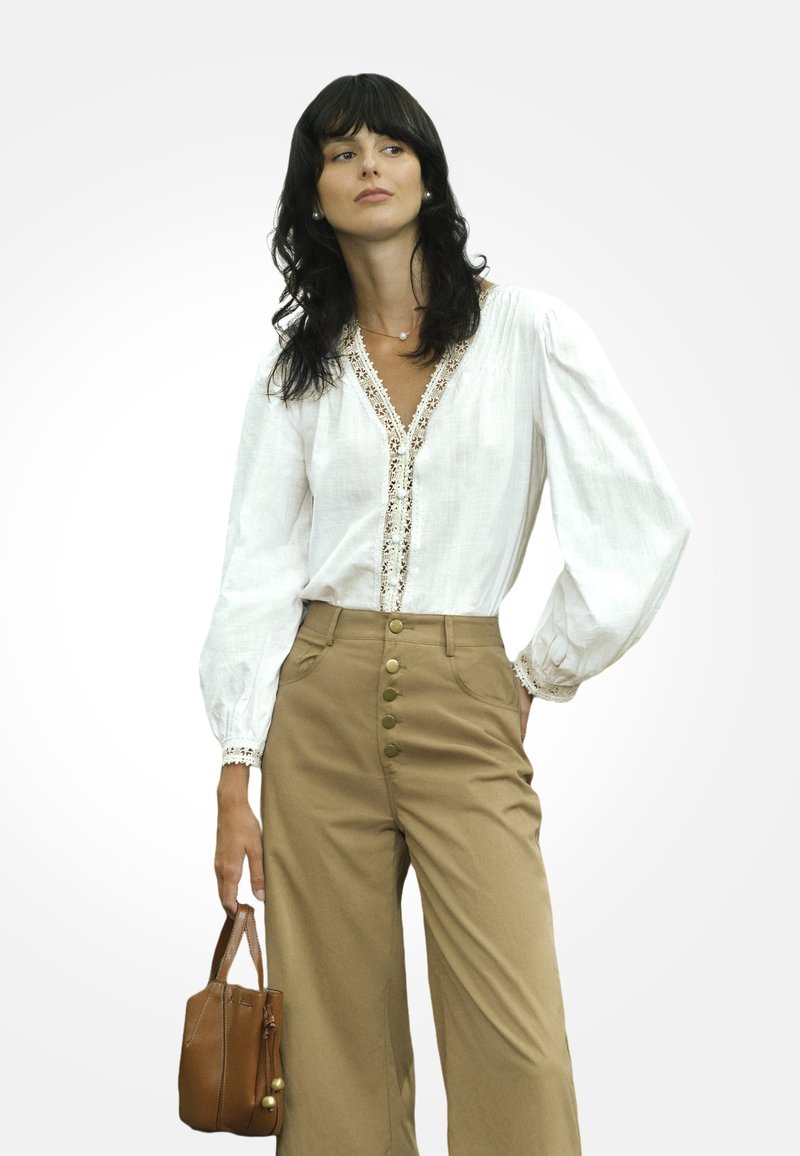 Mujer con cabello negro que lleva una blusa blanca con encaje, pantalones de tiro alto beiges con botones, sosteniendo un pequeño bolso marrón al costado.