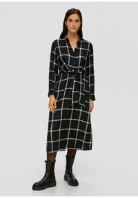 Schwarz-weiß karierte Hemdkleid mit langen Ärmeln, Taillenknoten und durchgehender Knopfleiste, kombiniert mit schwarzen Knöchelstiefeln.