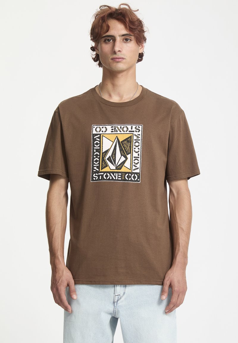 Brun bomulds t-shirt med korte ærmer og rund hals. Har et iøjnefaldende grafisk print i sort, hvid og gul på fronten.