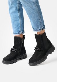 Bottines en suède noir avec une semelle épaisse, une fermeture éclair sur le côté et un accent en chaîne décoratif. Portées avec un jean bleu clair retroussé.