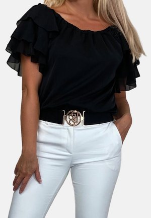 Mujer con blusa negra de mangas cortas con volantes, metida en pantalones blancos, con un cinturón negro que tiene una hebilla circular dorada.