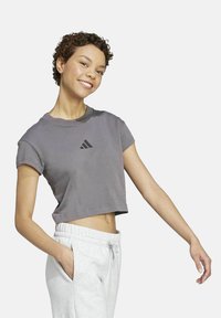 Gekruiste grijze t-shirt met korte mouwen en een zwart Adidas-logo op de borst, gecombineerd met lichtgrijze joggers. Gladde stofstructuur.