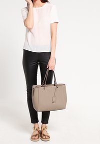 Gray tote bag with a structured shape, smooth texture, and dual handles. Worn with black pants and gold sandals. Simple, functional design.