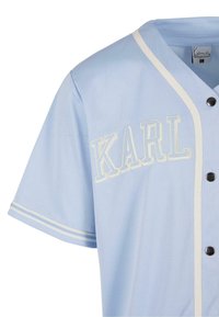 Maglietta a maniche corte azzurro chiaro con ricamo bianco "KARL" sul petto, bottoni neri e dettagli a strisce bianche sulle maniche.