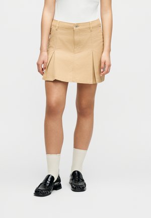 Γυναίκα που φοράει μπεζ πιε ντε πουλ μίνι φούστα με λευκό τοπ, λευκές κάλτσες και μαύρα παπούτσια λάκα, στέκεται μπροστά σε ένα απλό φόντο.