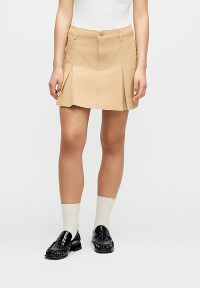 Femme portant une mini-jupe plissée beige avec un haut blanc, des chaussettes blanches et des mocassins en cuir verni noir, debout devant un fond uni.