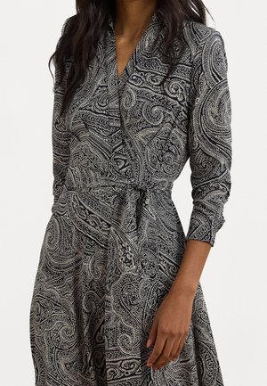 Femme portant une robe portefeuille à manches longues noire et blanche à motif cachemire avec une ceinture nouée à la taille sur un fond uni.