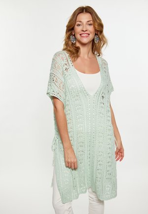 Helles mintgrünes Häkel-Überkleid mit tiefem V-Ausschnitt, das mit Spitzenmustern, seitlichen Bindebändern und kurzen Ärmeln versehen ist, über einem weißen Oberteil getragen.