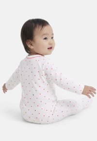 Νήπιο που φοράει λευκή πιτζάμα με κόκκινα σχέδια καρδιών, καθισμένο στο πάτωμα και κοιτάζει πλάγια με ένα ελαφρύ χαμόγελο.