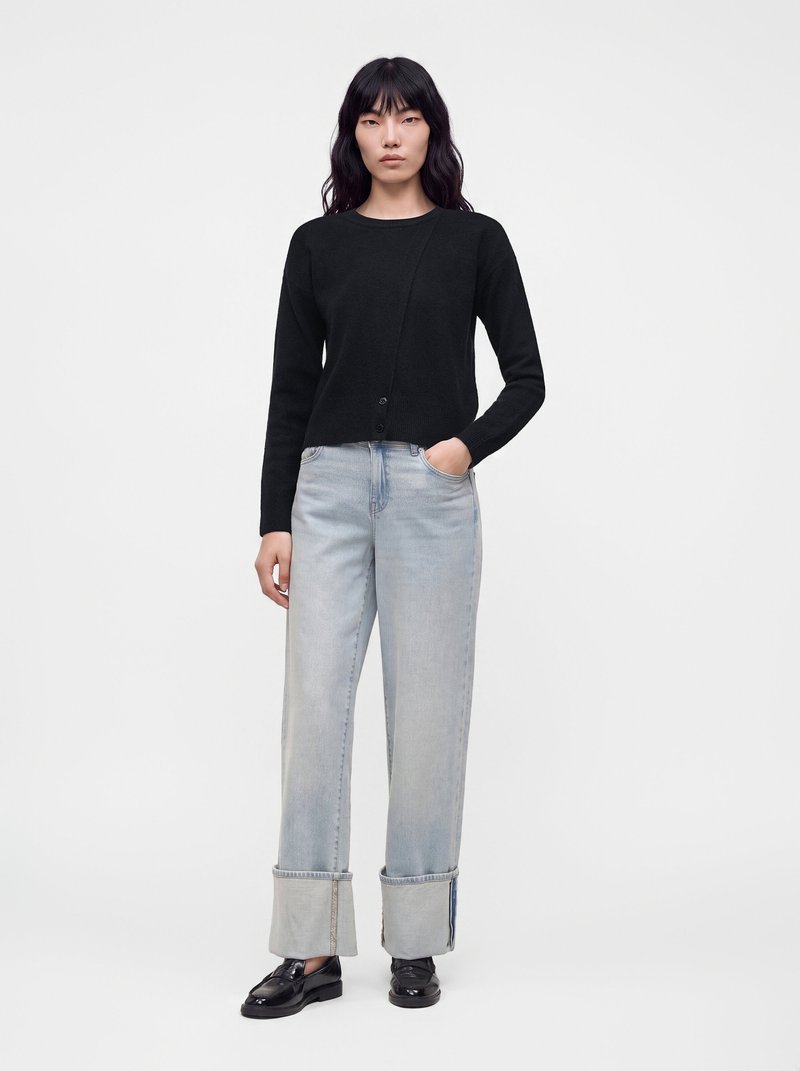 Femme aux longs cheveux noirs porte un pull noir, un jean large bleu clair avec de grands revers et des mocassins noirs brillants, debout sur fond blanc.