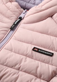 Jasnoróżowa kurtka pikowana z zamkiem błyskawicznym z przodu, czarnym logo "SUPERDRY" oraz teksturowanym materiałem z przeszyciami w kształcie poduszek i fioletowym akcentem.