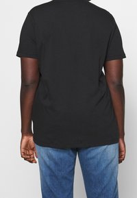 Zwarte katoenen t-shirt met korte mouwen, ronde halslijn en een gladde structuur. Draagt met blauwe denim jeans, gezien van achteren.