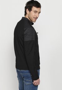 Jachetă neagră cu material texturat și accente negre lucioase la umeri. Croială slim fit, cu guler și buzunare laterale. Asortată cu blugi albaștri.
