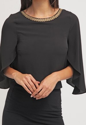 Žena v černé blůze se širokými, volnými rukávy a zlatým řetízkovým ozdobením podél výstřihu, ruce jemně sepnuté v pase.