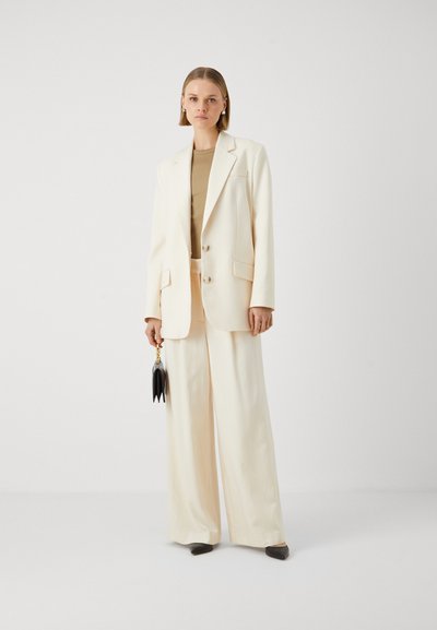 Blazer crema con vestibilità tailored, due bottoni e tasche laterali, abbinato a pantaloni a gamba larga. Gli accessori includono una borsa nera.