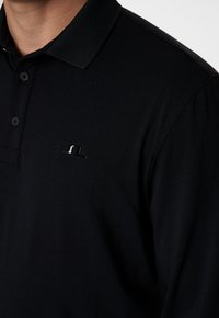Nahaufnahme einer Person, die ein schwarzes Poloshirt mit strukturiertem Kragen und einem kleinen glänzenden Logo auf der linken Brust trägt.