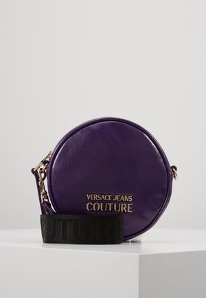 Okrągła fioletowa torebka na ramię Versace Jeans Couture ze złotym logo i szerokim czarnym regulowanym paskiem na białej powierzchni.