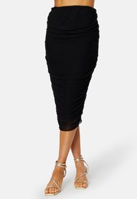 Czarna spódnica sięgająca do kolan, z marszczonym wzorem i pionowymi plisami. Materiały wydają się gładkie, o dopasowanym kształcie. Założona z złotymi sandałami na obcasie.