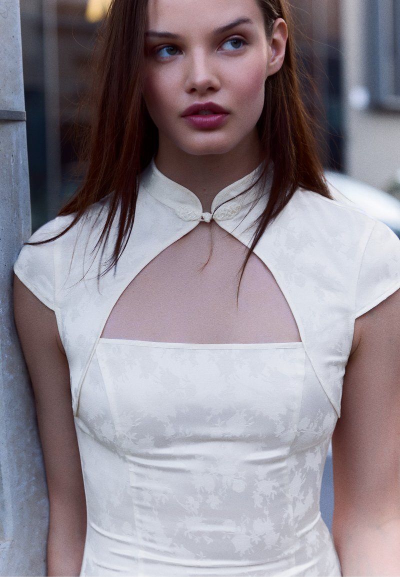 Jeune femme aux longs cheveux bruns portant une robe en satin blanc avec un col haut et un détail découpé au niveau de la poitrine, debout à l'extérieur.
