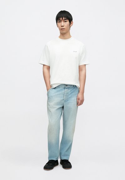 Άσπρο βαμβακερό μπλουζάκι με μικρό μαύρο λογότυπο, σε συνδυασμό με ανοιχτό μπλε τζιν παντελόνι και μαύρα αθλητικά παπούτσια, στέκεται σε απλό φόντο.