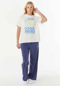Camiseta blanca con un gráfico colorido del hotel Aloha, combinada con pantalones de pierna ancha en azul marino. Ambas prendas tienen un corte holgado y texturas suaves.