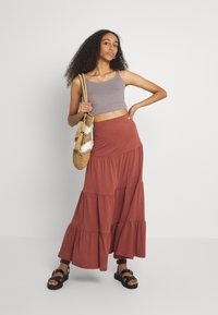 Un crop top gris et une jupe maxi tiered couleur rouille, tous deux en tissu doux. Le modèle tient un sac à main tressé et porte des sandales noires.