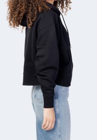 Czarna, krótka bluza z kapturem, z kieszenią z przodu, sznurkiem do regulacji kaptura i ściągaczami przy rękawach, noszona z jasnoniebieskimi spodniami dżinsowymi o wysokim stanie.