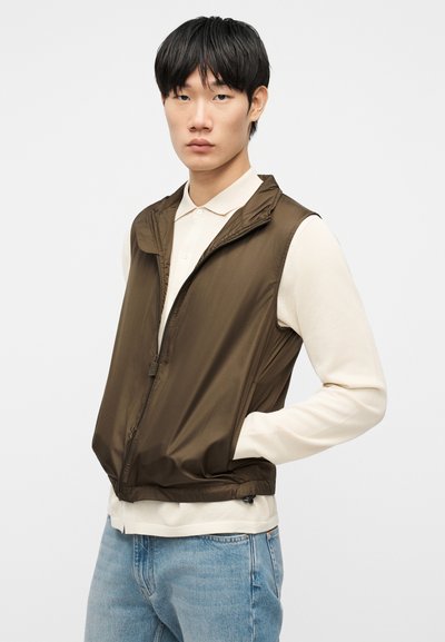 Νεαρός άνδρας με μαύρα μαλλιά, που φοράει καφέ γιλέκο με φερμουάρ πάνω από κρεμ μακρυμάνικο πουκάμισο και ανοιχτόχρωμο τζιν, στέκεται μπροστά σε απλό φόντο.