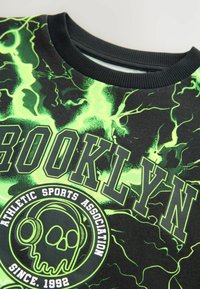 Schwarzer Pullover mit einem leuchtend grünen Blitzmuster. "BROOKLYN" und ein Totenkopf-Graphic mit Kopfhörern in kräftigem Schwarz und Weiß gedruckt.