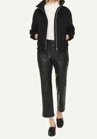 Kvinna klädd i en svart zip-up jacka, vit topp, svarta läderbyxor och svarta slip-on skor som står med händerna i fickorna.