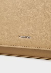 Cuir texturé tan avec un grain subtil. Présente un logo « Calvin Klein » en métal argenté embossé sur la surface, des lignes épurées et aucun matériel visible.