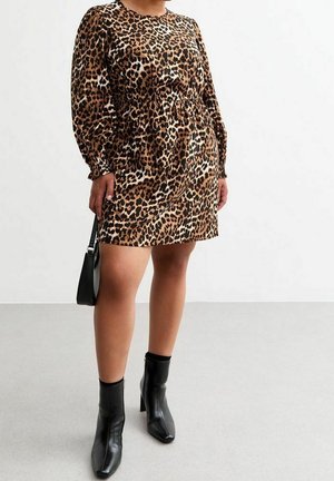 Frau trägt ein kurzes Kleid mit langen Ärmeln und Leopardenmuster, hält eine schwarze Handtasche und trägt schwarze Stiefeletten mit blockabsätzen.