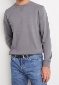 Felpa grigia a maniche lunghe con collo a giro e piccolo logo sul petto, abbinata a jeans in denim blu e una cintura nera.