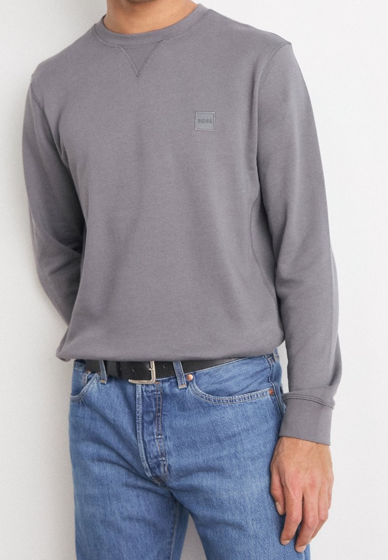 Felpa grigia a maniche lunghe con collo a giro e piccolo logo sul petto, abbinata a jeans in denim blu e una cintura nera.