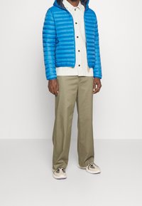 Giaccone puffer blu con strisce orizzontali, con cappuccio, sopra una maglietta a righe bianche. Pantaloni verde oliva e sneaker chiare completano l'outfit.