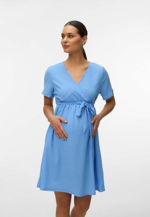 Mujer embarazada con cabello oscuro recogido, vistiendo un vestido cruzado de manga corta color azul claro, con las manos apoyadas suavemente sobre su vientre.