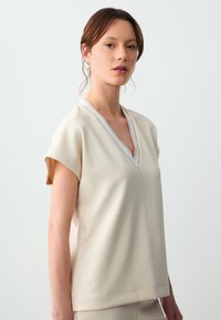 Beige kort mouwtop met een V-hals en contrasterende afwerking. Zachte textuur, ontspannen pasvorm en minimalistisch ontwerp.