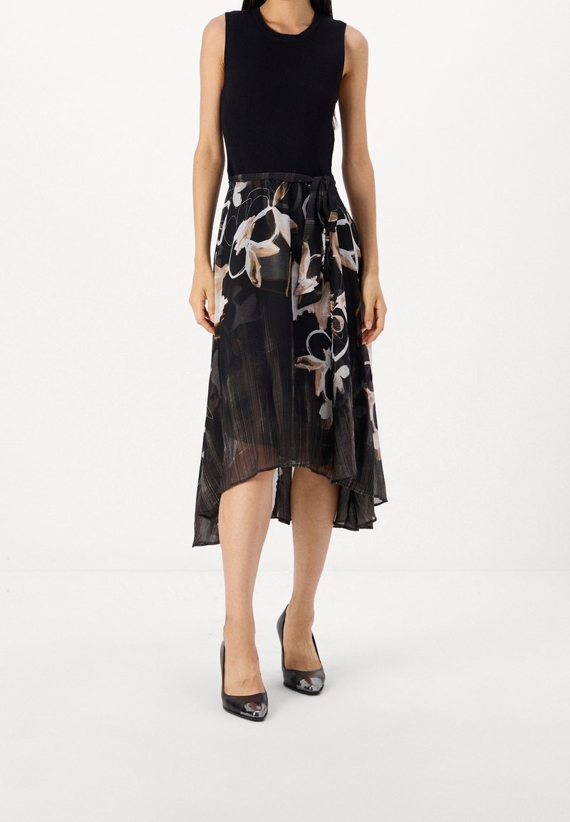 Un haut sans manches noir et une jupe fluide de longueur genoux avec un motif floral dans des teintes noires, crème et brunes ; assortis à des chaussures à talons noires.